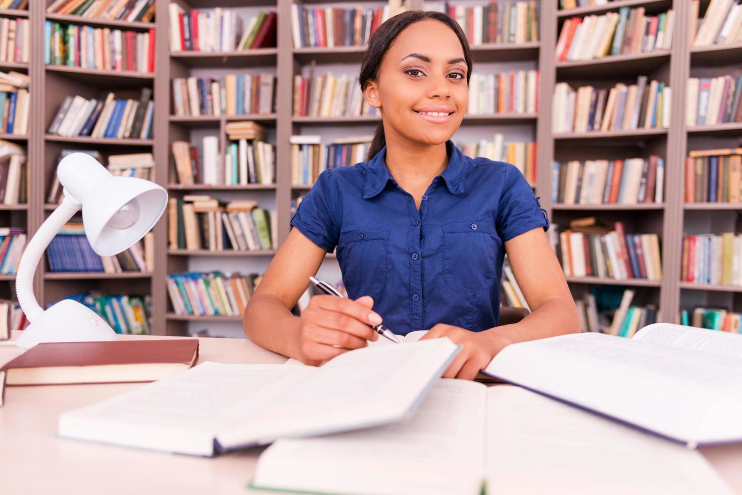 framställning till henne slutlig tentor. självsäker ung svart kvinna ser på kamera och leende medan Sammanträde på de bibliotek skrivbord foto