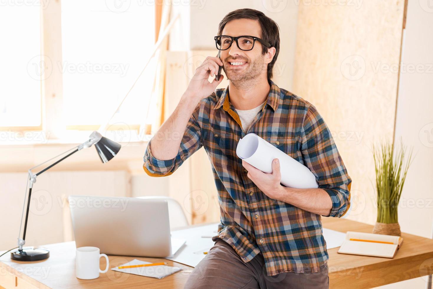 excellent resultat av hans arbete. Lycklig ung man innehav plan och talande på de mobil telefon medan lutande på hans arbetssätt plats foto