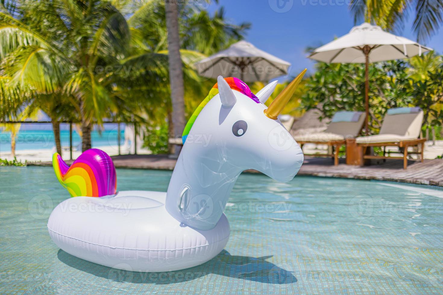 uppblåsbar färgrik vit enhörning på tillflykt simning slå samman. semester tid i tropisk slå samman med plast leksaker. avslappning och roligt begrepp, panorama- sommar bakgrund. handflatan träd, stolar, sängar paraplyer foto