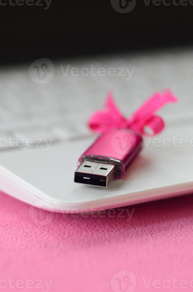 lysande rosa uSB blixt minne kort med en rosa rosett lögner på en filt av mjuk och hårig ljus rosa skinna tyg bredvid till en vit bärbar dator. klassisk kvinna gåva design för en minne kort foto