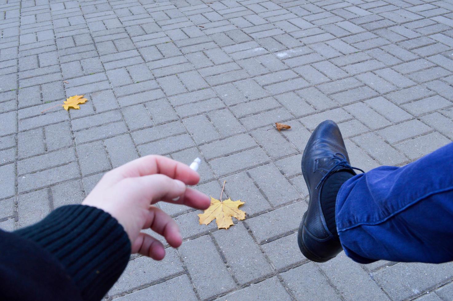 en man innehar en cigarett i hans händer, fingrar Sammanträde i de parkera och rökning med hans ben i en sko mot de bakgrund av stenläggning plattor med höst löv. de bakgrund foto