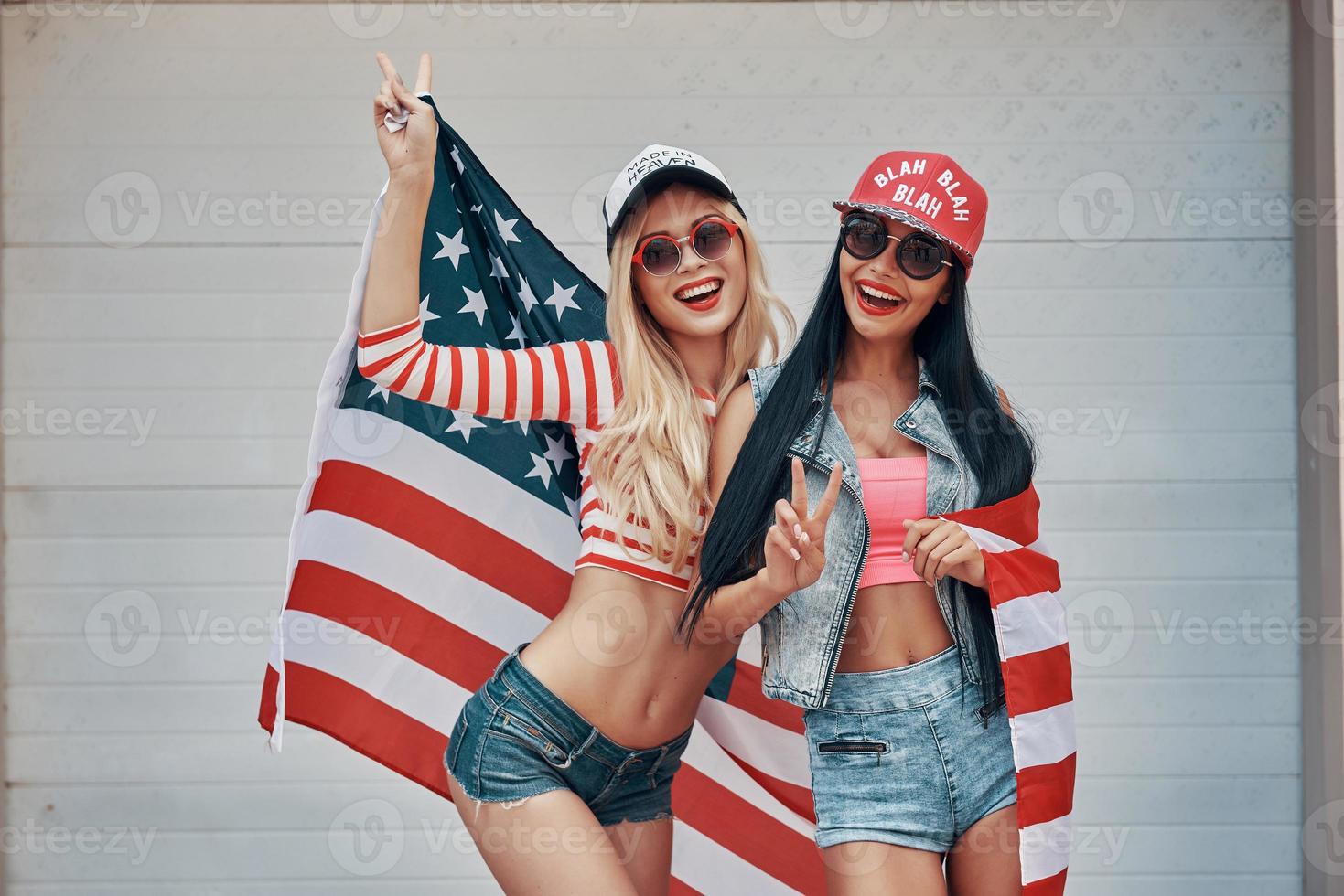 amerikan flickor. två lekfull ung kvinnor gestikulerar fred tecken och innehav amerikan flagga medan stående mot de garage dörr foto