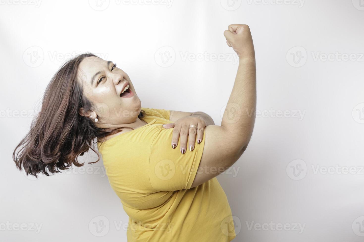 upphetsad asiatisk stor sized kvinna bär en gul t-shirt som visar stark gest förbi lyft henne vapen och muskler leende stolt foto