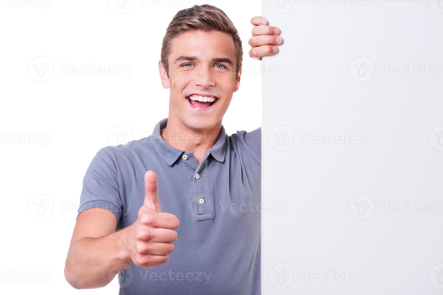 tumme upp Lycklig ung man ser på kamera och som visar hans tumme upp medan lutande på de kopia Plats och stående mot vit bakgrund foto