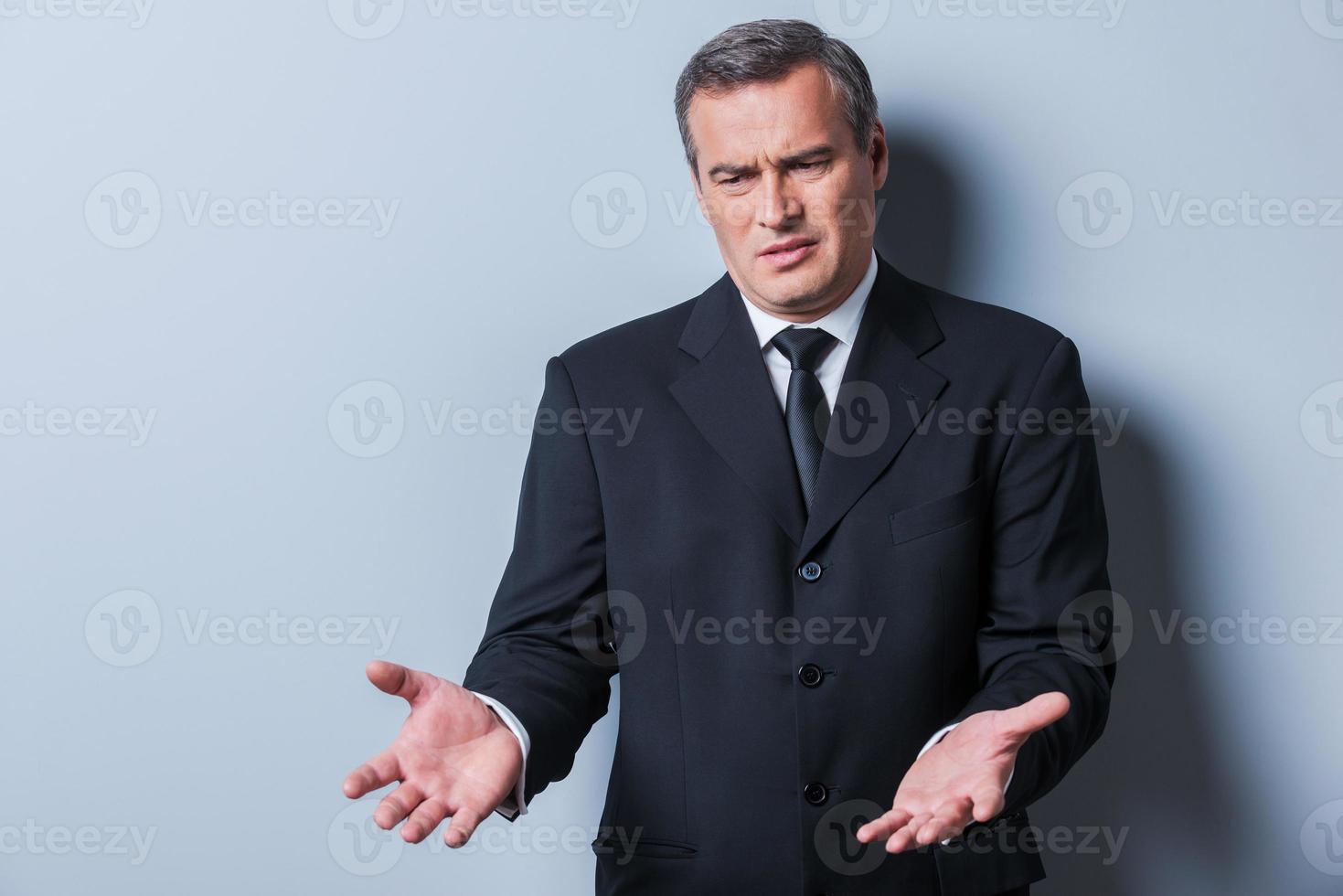Vad är den där missnöjd mogna man i formell klädsel gestikulerar och ser bort medan stående mot grå bakgrund foto