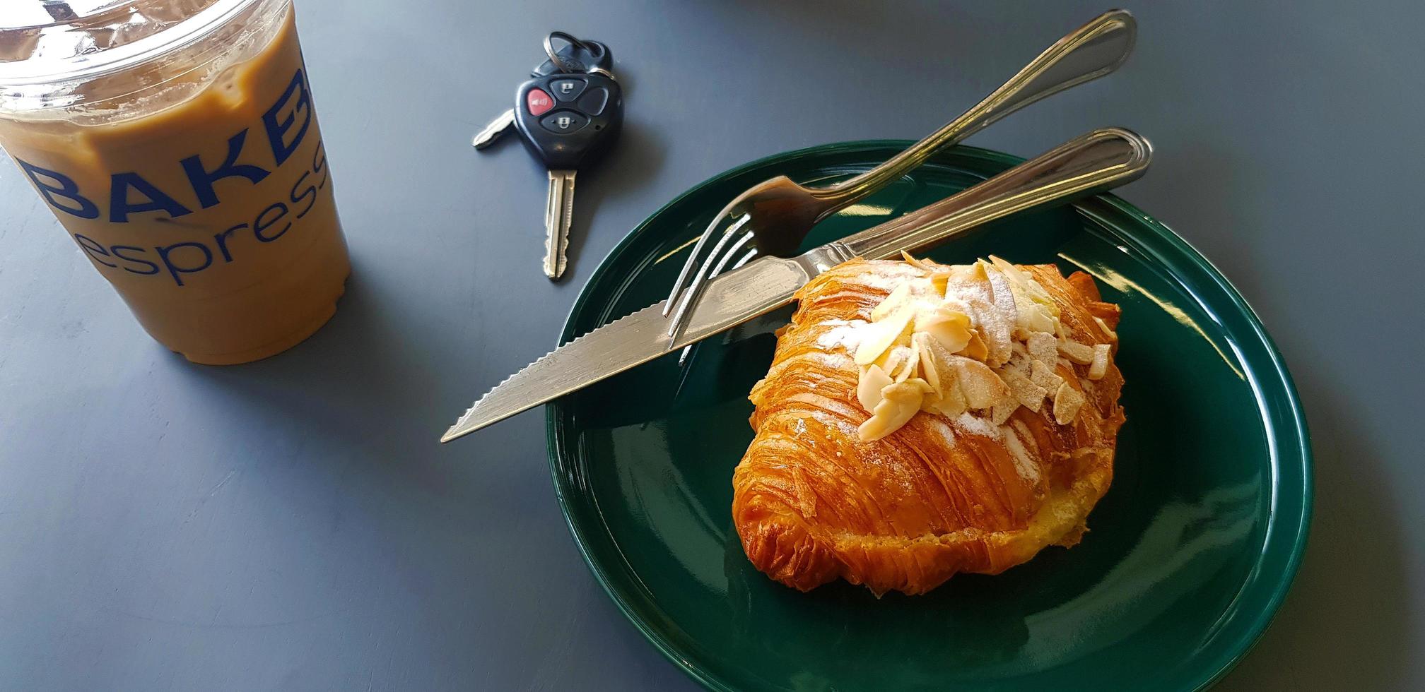 croissant med glasyr socker och mandel skiva med bil avlägsen nyckel, iced espresso kaffe rostfri stål kniv i grön maträtt på grå tabell med kopia Plats. Krispig bröd på tallrik på grå tabell bakgrund. foto