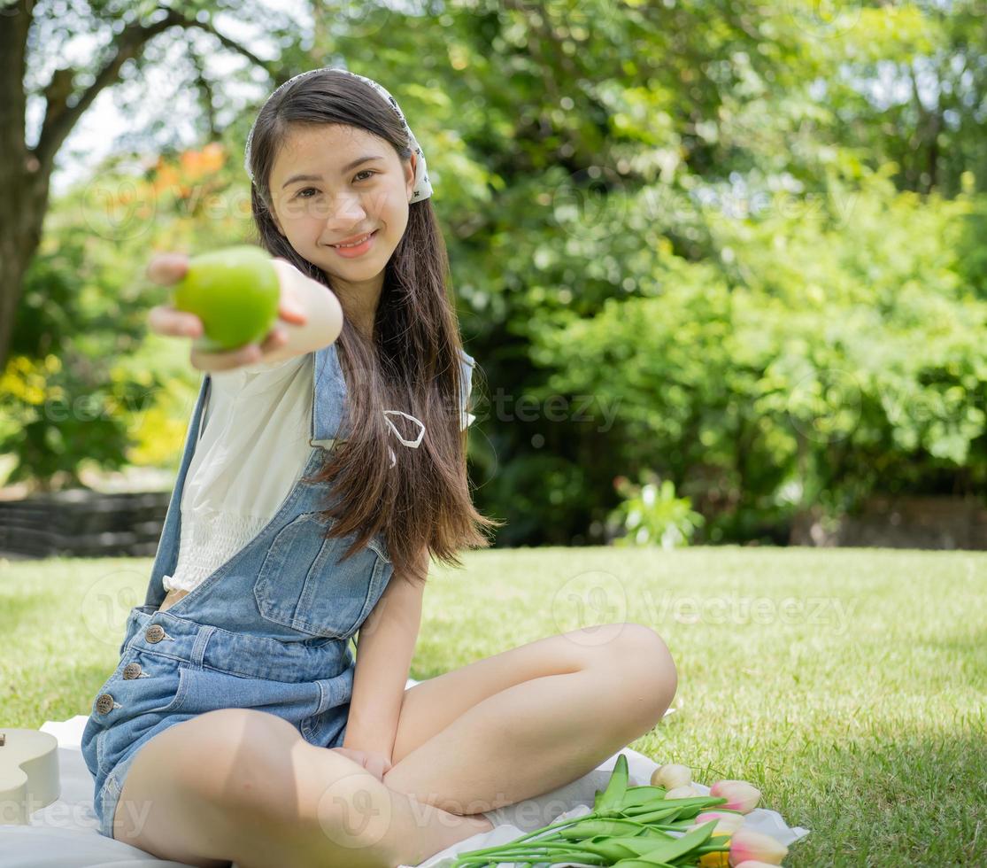 ung flicka person innehav äpple frukt, färsk vitamin diet, friska livsstil, porträtt. glad skön asiatisk Tonårs Lycklig Sammanträde utomhus- fritid aktivitet i grön parkera. leende barn spela äpple foto