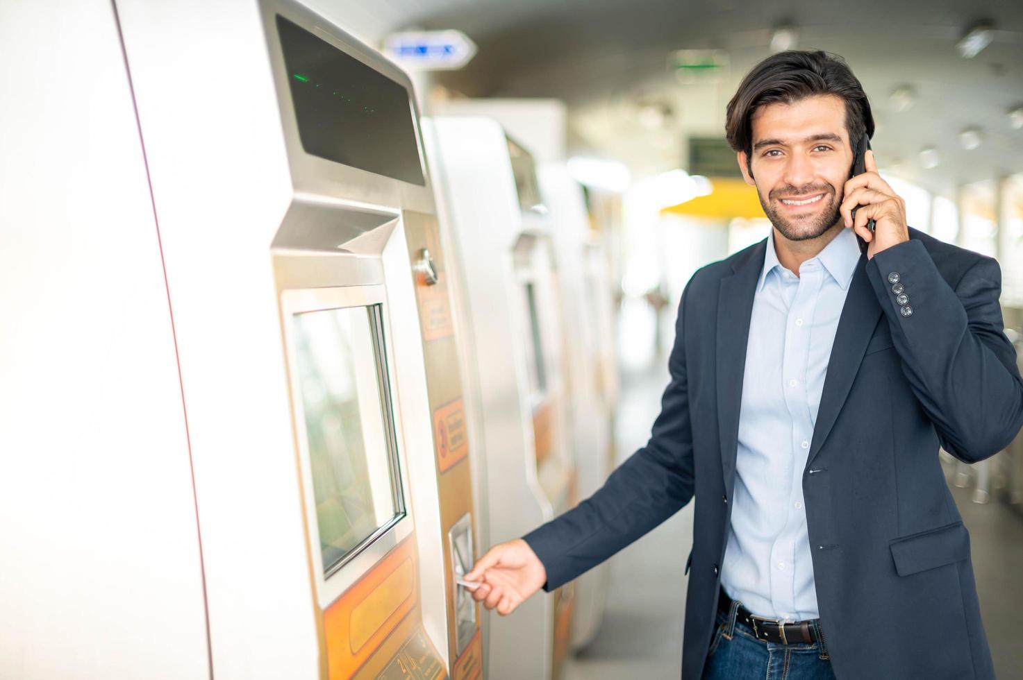 de caucasian man använda sig av en transport kort på de biljett maskin för resa förbi himmel tåg i de rusa timme tid medan han användningar smartphone för talande med hans vän. foto