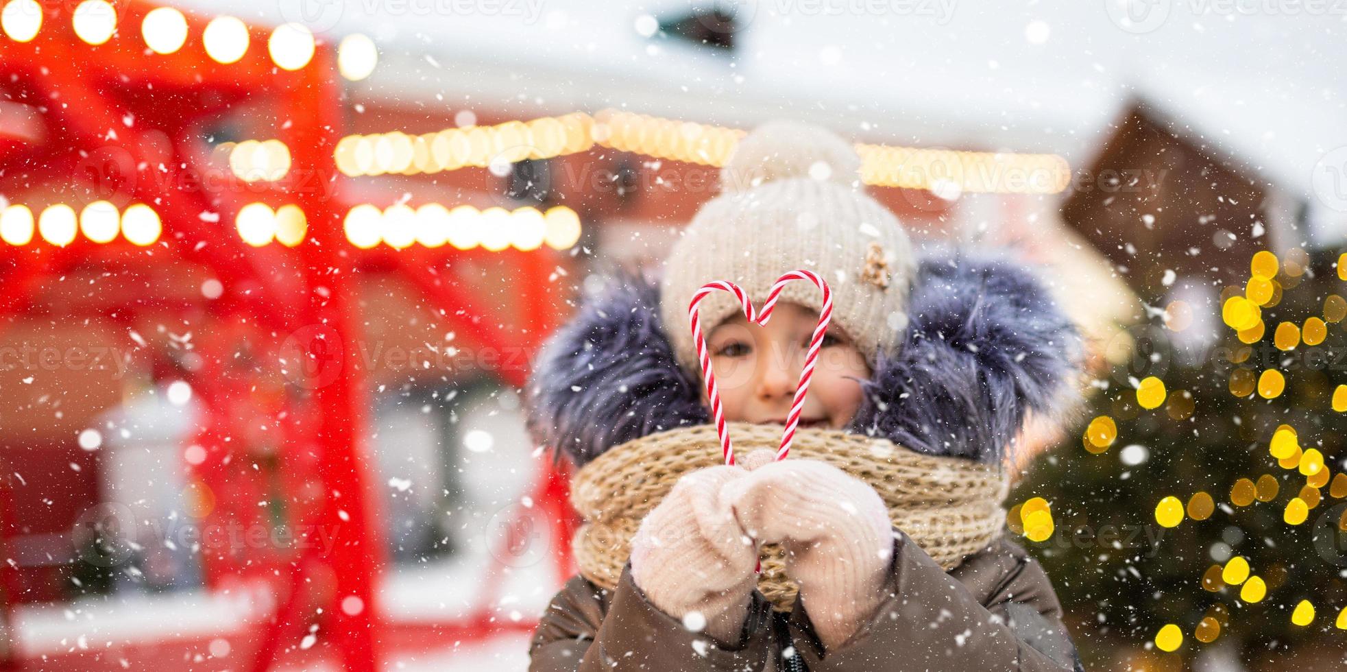 Söt flicka innehar i hans händer godis sockerrör i de form av en hjärta utomhus- i värma kläder i vinter- festlig marknadsföra. fe- lampor kransar dekorerad snö stad för ny år. jul humör foto