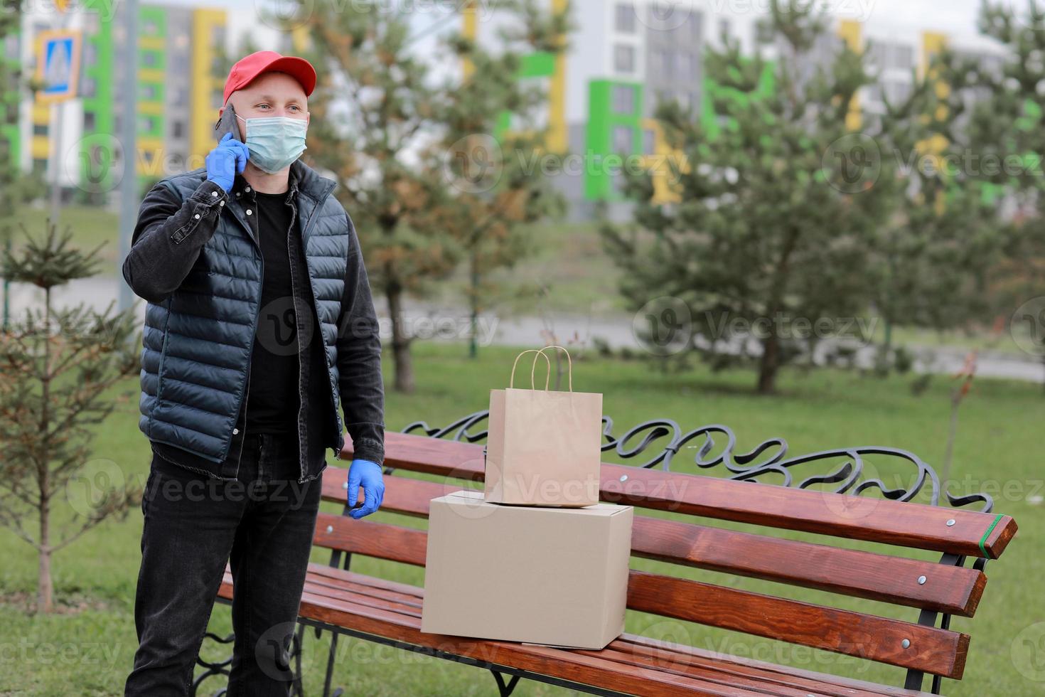 leverans man i röd hatt, skydd mask och medicinsk sudd handskar med skiften använder sig av smartphone till sändning till kund. leverans service foto