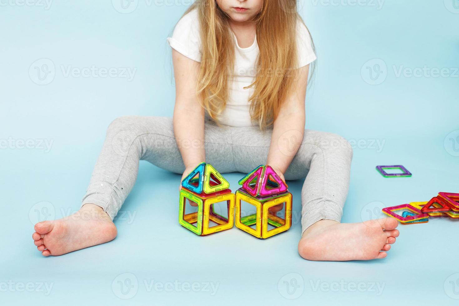 liten flicka spelar med färgrik magnetisk konstruktör leksak på blå bakgrund. barn byggd en hus foto