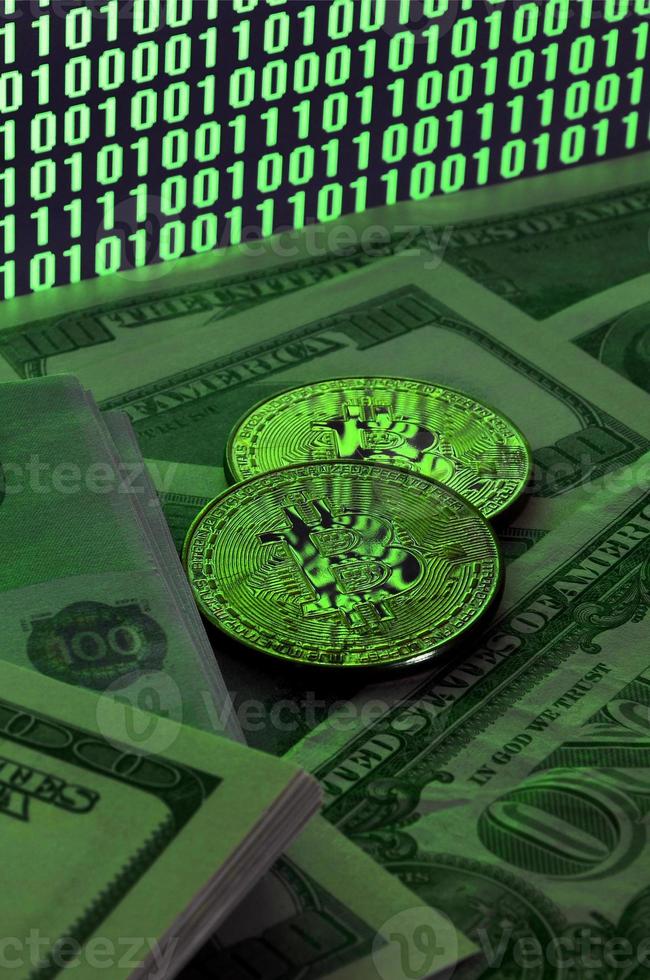 två bitcoins lögner på en lugg av dollar räkningar på de bakgrund av en övervaka skildrar en binär koda av ljus grön nollor och ett enheter på en svart bakgrund. låg nyckel belysning foto