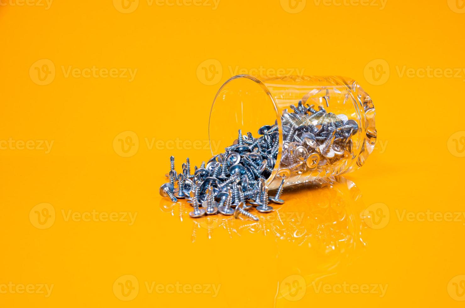 många metall självuttag skruvar tillverkad av stål i en glas, i hink burk. självuttag skruva för metall, för järn, förkromad självuttag skruva, på ett orange gul bakgrund, foto