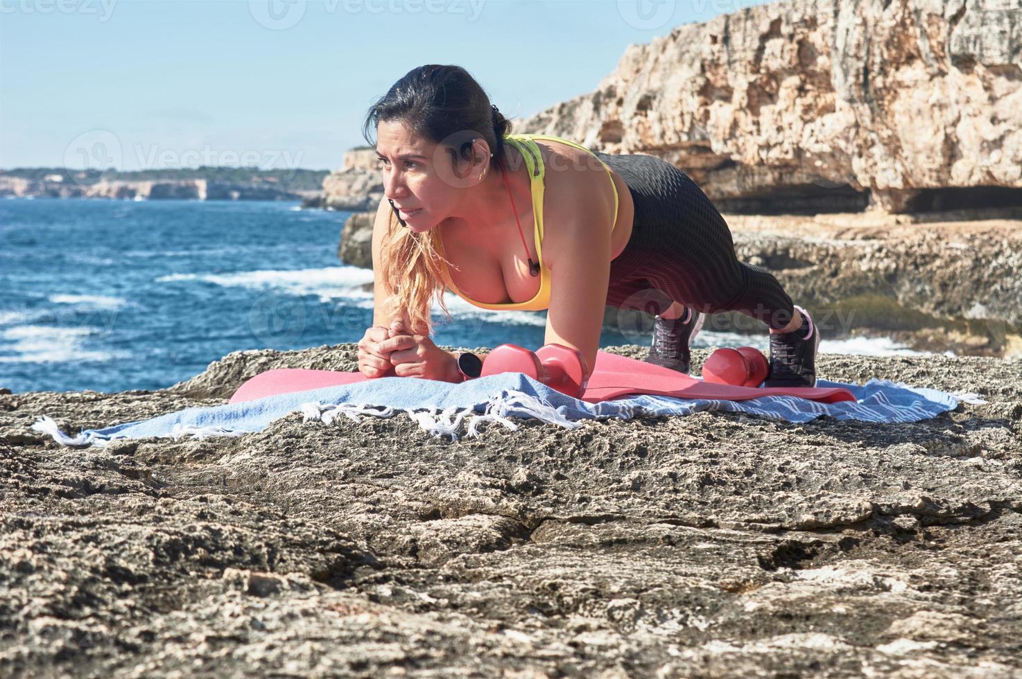 latin kvinna, medelålders, bär sportkläder, Träning, håller på med fysisk övningar, planka, situps, klättrarens steg, brinnande kalorier, förvaring passa, utomhus förbi de hav, bär hörlurar, smart Kolla på foto