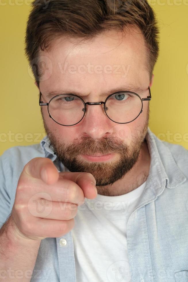 man med en skägg och glasögon strikt utseende och poäng fingrar, på en gul bakgrund. foto