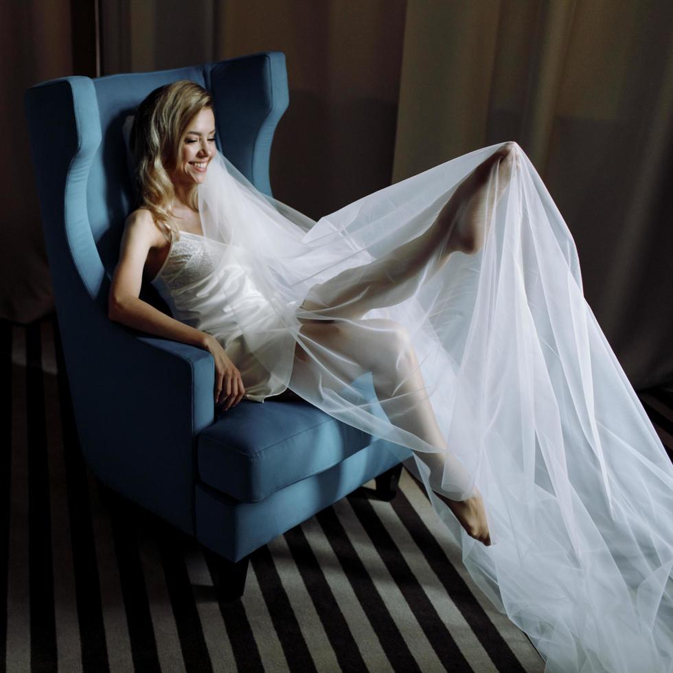 bruden lyfter upp benen och sitter i en stor blå stol i hotellrummet foto