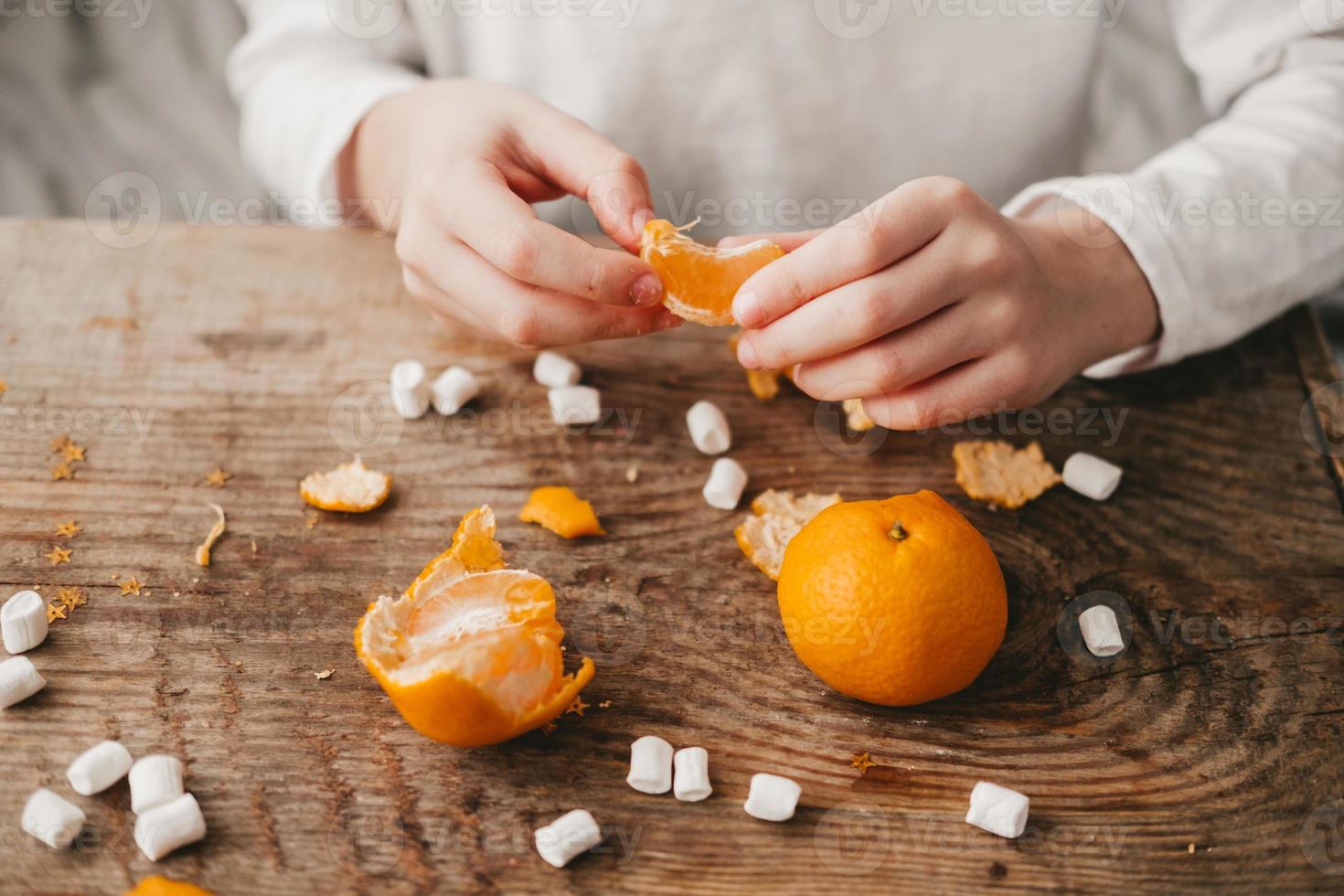 händer av en barn rengöring en mandarin på en trä- bakgrund, topp se. jul humör. orange frukt med marshmallows. frukt med vitamin c. sötsaker för de ny år foto