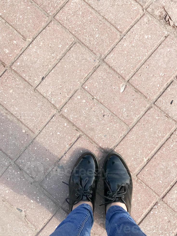 topp se av de ben, skor, flickor fötter, kvinnor i patent läder skor på de bakgrund av en sten brun röd betong fyrkant stenläggning rektangulär bricka foto