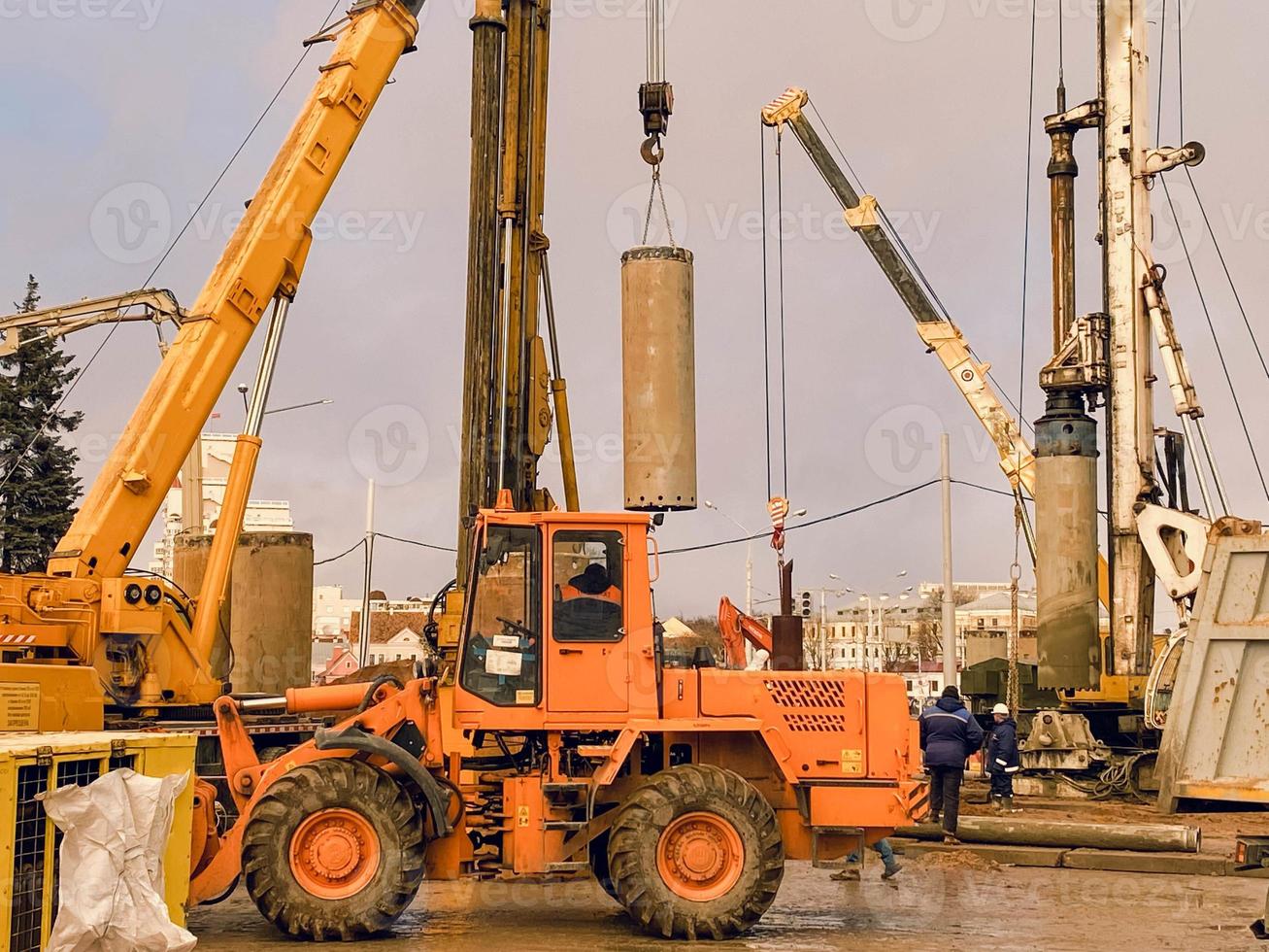 konstruktion Utrustning på de planskild reparera webbplats. stor betong block är restes förbi kranar. ett orange traktor för asfalt stenläggning enheter närliggande på stor hjul foto