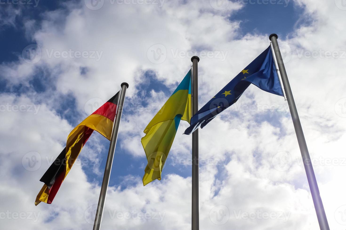 Ukrainas, Tysklands och Europeiska unionens flaggor flyger sida vid sida mot blå himmel. foto