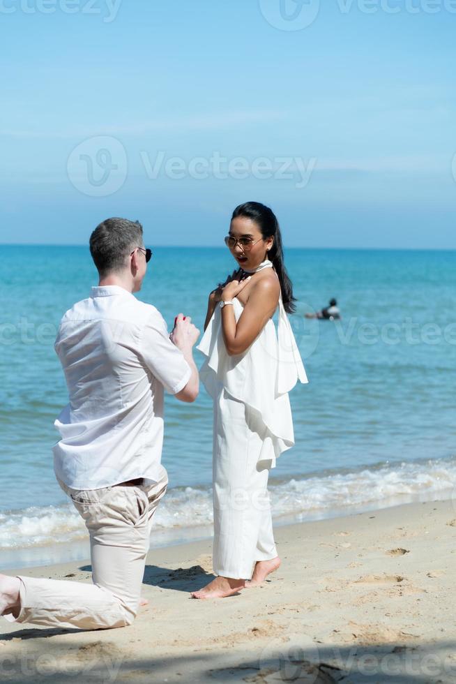 en ung vit man be för ett asiatisk flicka till gifta sig med en diamant ringa på en strand foto