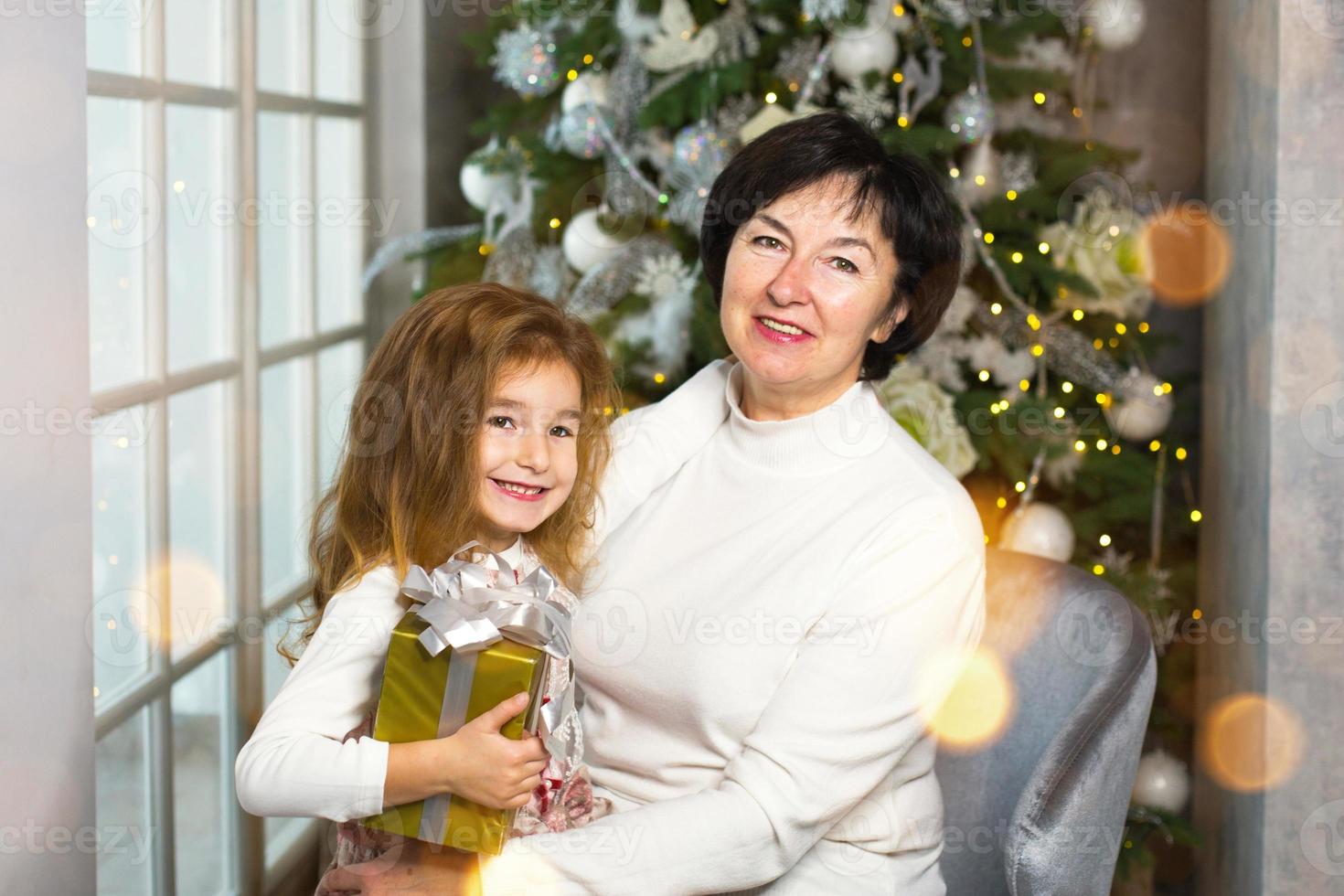mormor med en liten flicka på de bakgrund av jul dekorationer och en stor fönster. familj Semester, känslor, gåva låda. barnbarn på mormors knä. ny år foto