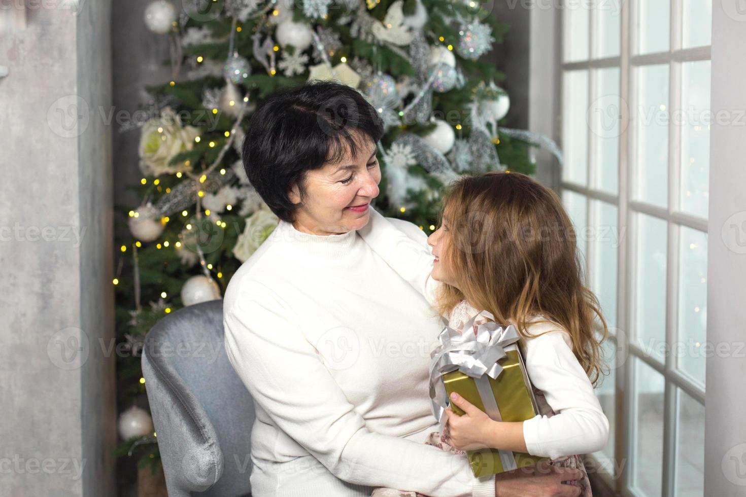 mormor med en liten flicka på de bakgrund av jul dekorationer och en stor fönster. familj Semester, känslor, gåva låda. barnbarn på mormors knä. ny år foto