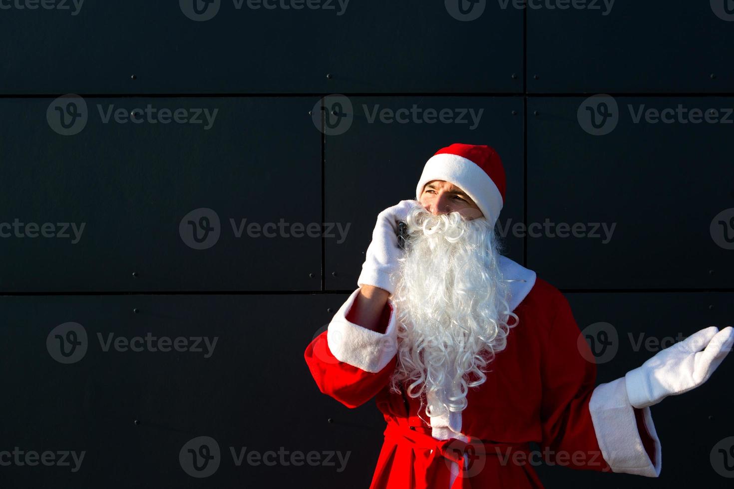 modern santa claus talande på de telefon. beställning de tjänster av ett animatör för de jul och ny år. uppkopplad hälsningar via de internet och mobil kommunikation, social distans foto