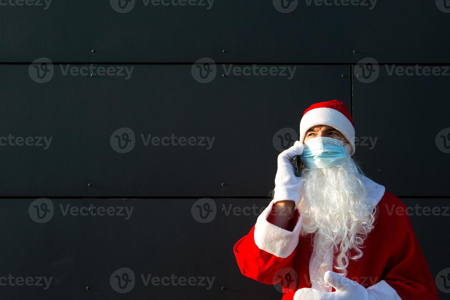 santa claus i en medicinsk mask med en smartphone. uppkopplad hälsningar, beställning tjänster för jul och ny år. social distans i de coronavirus epidemi, en ny verklighet. kopia Plats foto