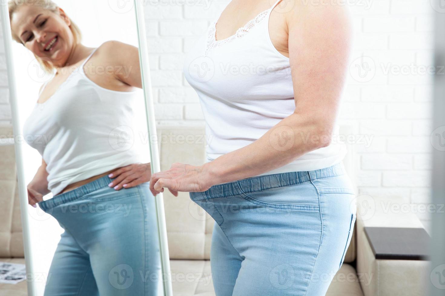 missnöjd mitten åldrig kvinna ser på själv i de spegel och klämmande mage fett i inhemsk interiör. ledsen plus storlek kvinna ser henne reflexion i de spegel med sorg på Hem. foto