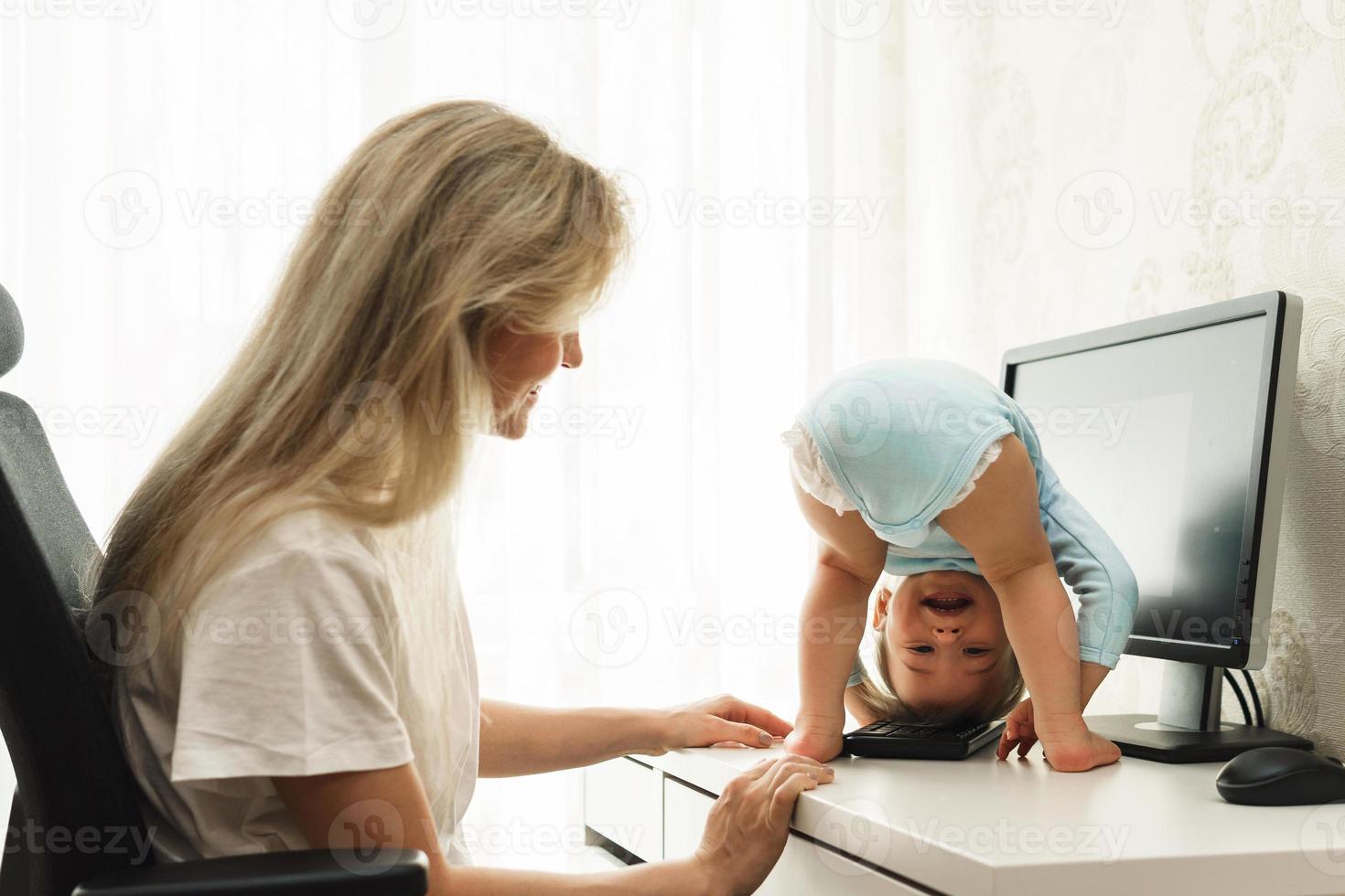 liten pojke stående upside ner på en skrivbord och distraherande mor från arbete på dator. foto