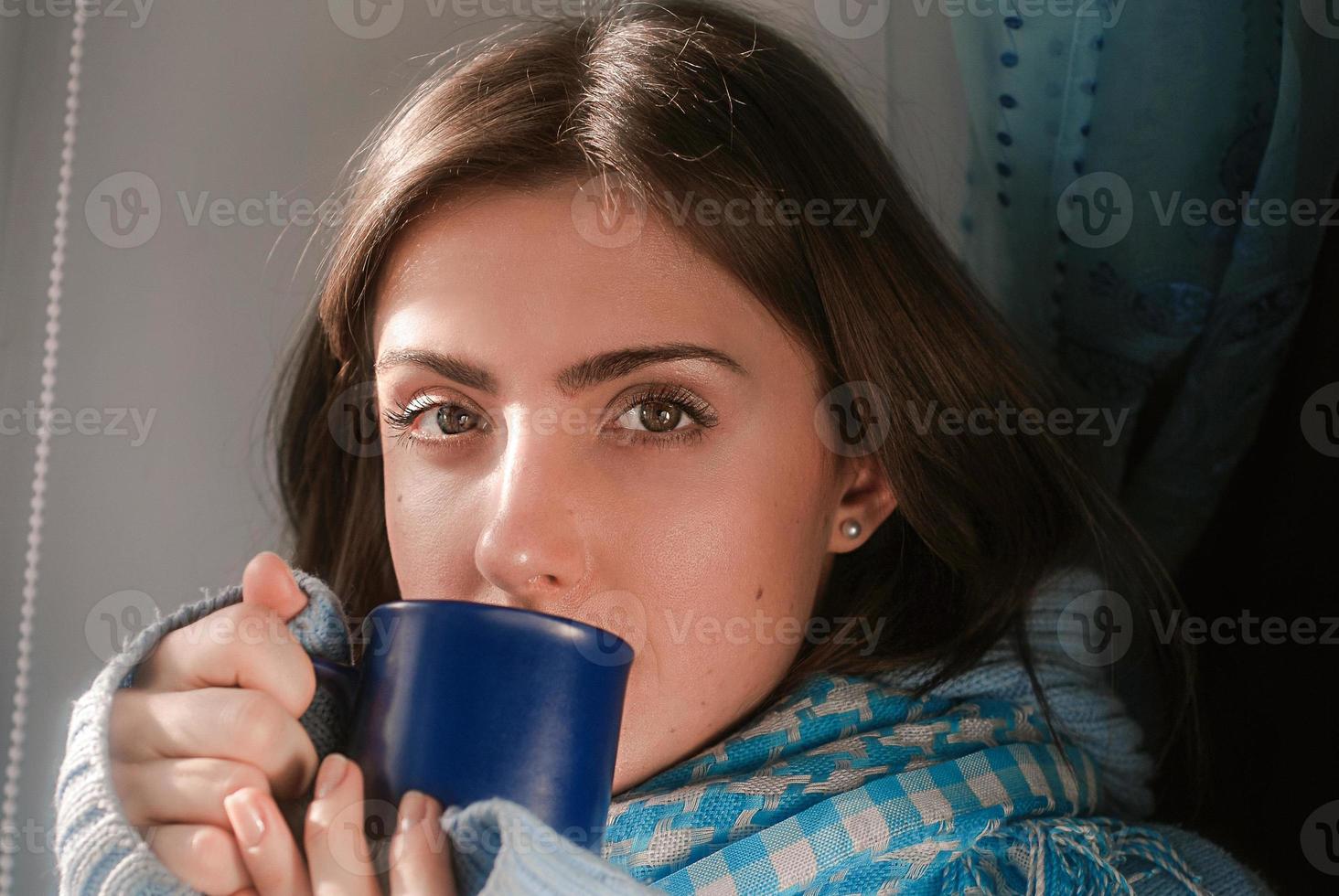 ledsen Söt ung kvinna vistas Hem eftersom av de influensa och temperatur i värma hoppare och scarf med kopp av te nära de fönster foto