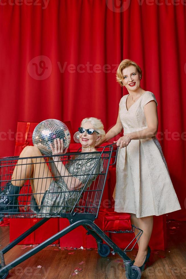 mogna kvinna rullande eleganta senior kvinna i solglasögon och silver- klänning de mataffär vagn på de fest. fest, disko, firande, senior ålder begrepp foto