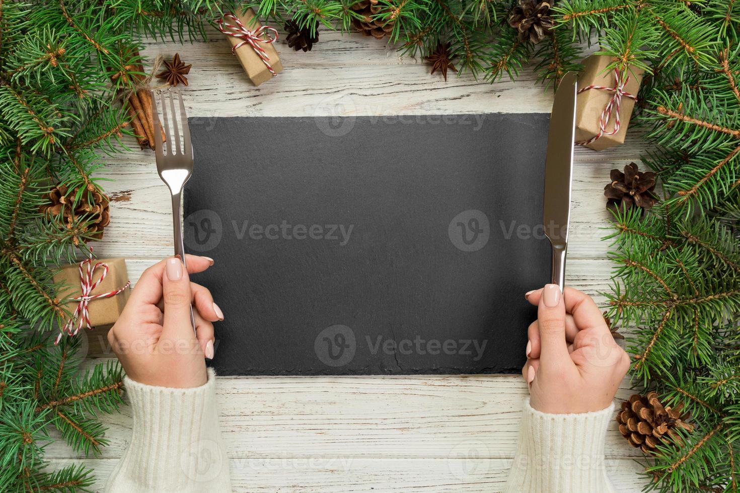 topp se flicka innehar gaffel och kniv i hand och är redo till äta. tömma svart skiffer rektangulär tallrik på trä- jul bakgrund. Semester middag maträtt begrepp med ny år dekor foto