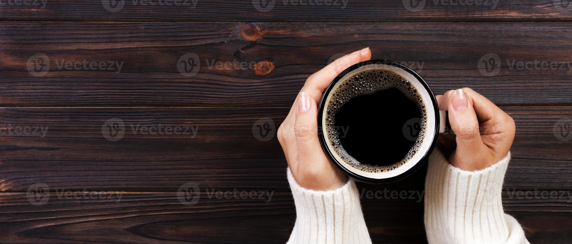ensam kvinna dricka kaffe i de morgon, topp se av kvinna händer innehav kopp av varm dryck på trä- skrivbord. baner foto