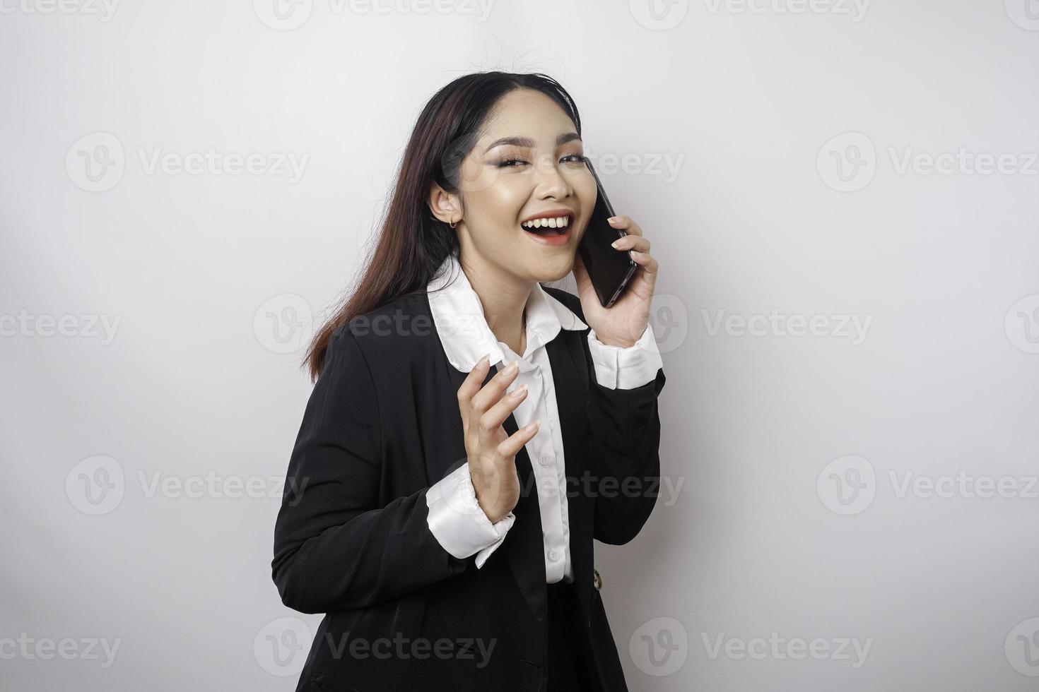 en porträtt av en Lycklig asiatisk affärskvinna är leende medan talande på telefon ring upp bär en svart kostym isolerat förbi en vit bakgrund foto