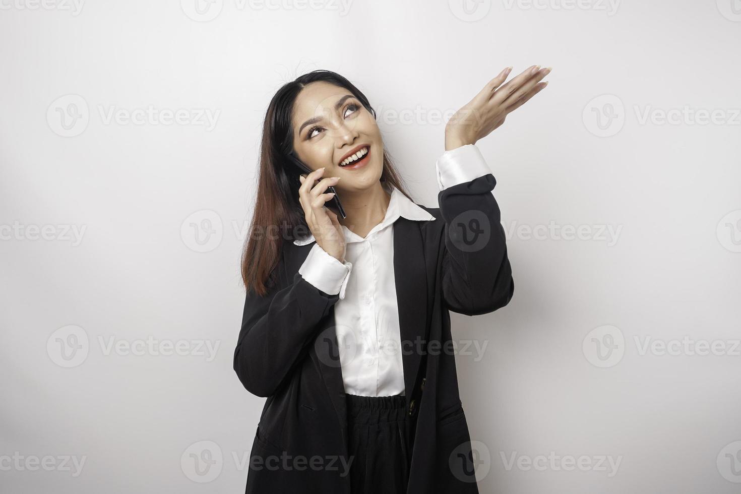 upphetsad asiatisk affärskvinna bär svart kostym pekande på de kopia Plats på topp av henne medan talande på en telefon ringa upp, isolerat förbi vit bakgrund foto