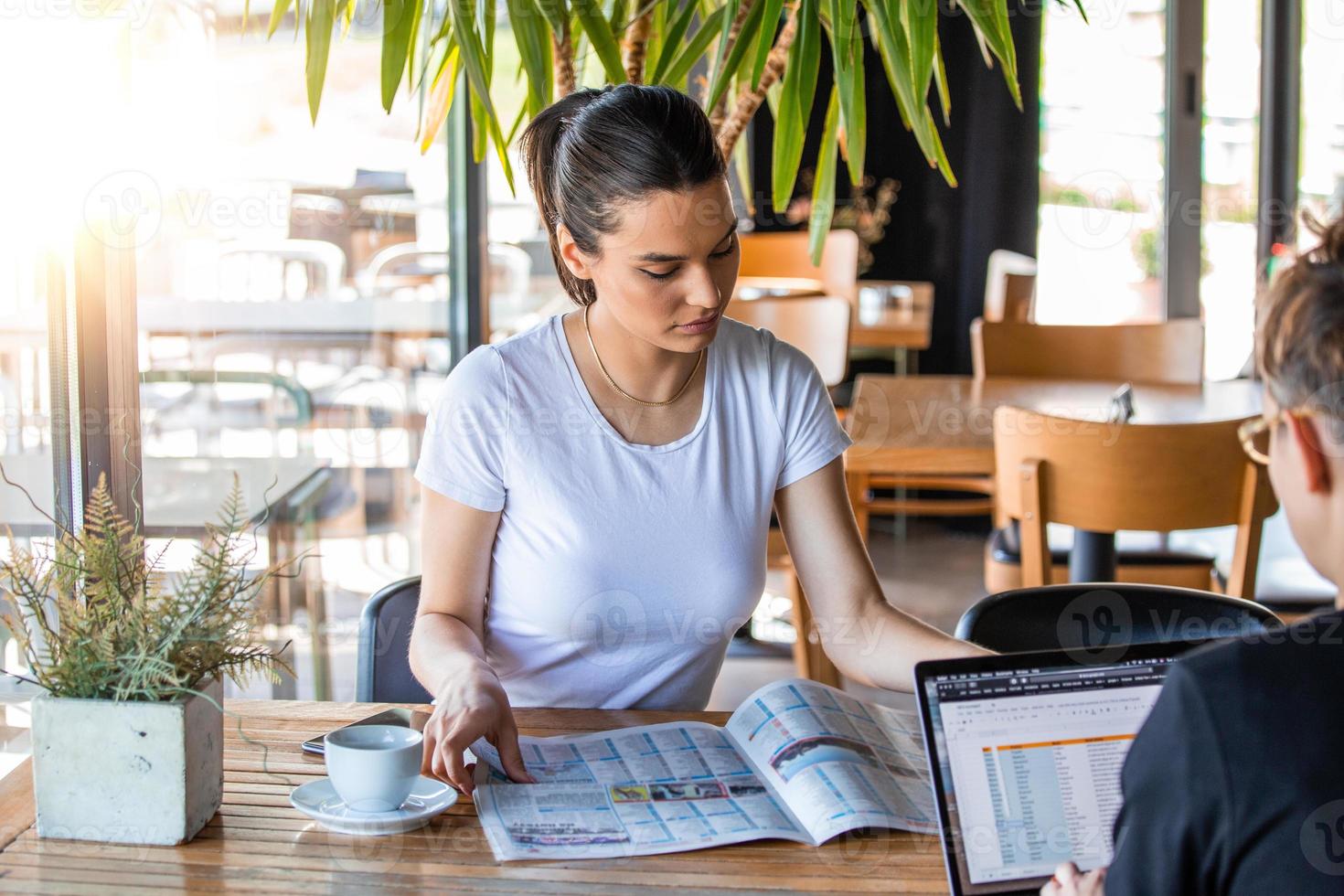 charmig kvinna med henne vän läsning tidning under resten i kaffe affär, Lycklig caucasian kvinna läsning Nyheter medan avkopplande i Kafé under fri tid foto