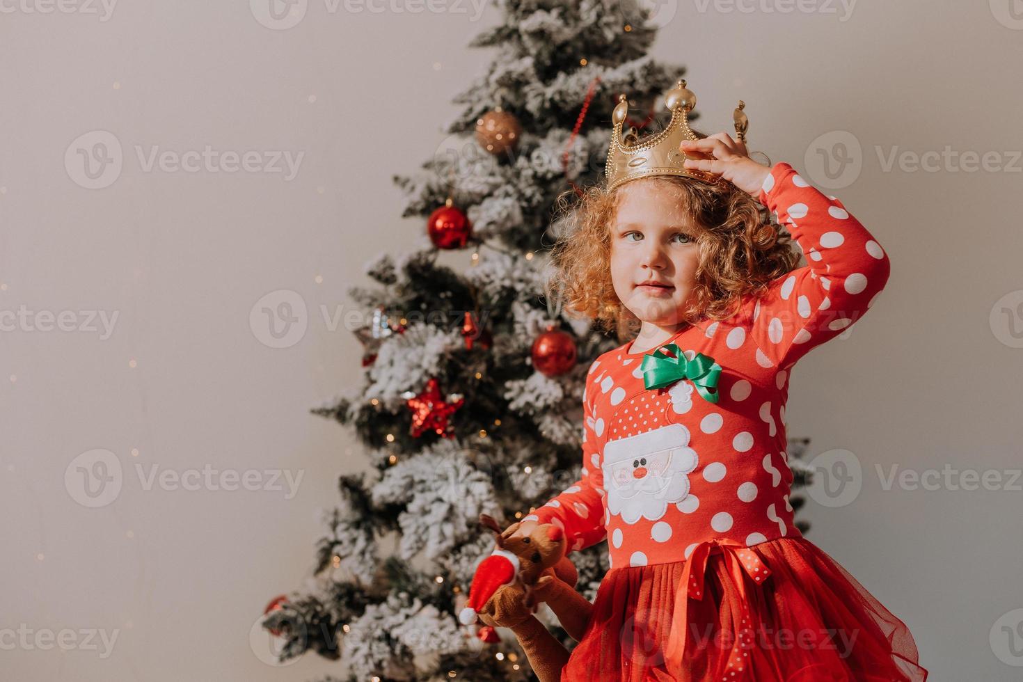 liten lockigt hår flicka i en karneval klänning gömde sig henne ansikte Bakom skinande jul träd leksaker stjärnor. barn i en röd klänning med en santa skriva ut på de bakgrund av en jul träd. hög kvalitet Foto