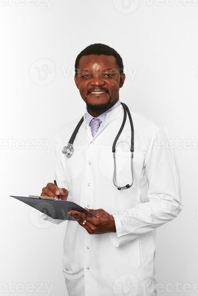 leende svart skäggig läkare man i vit morgonrock med stetoskop fyllning medicinsk uppgifter på Urklipp foto