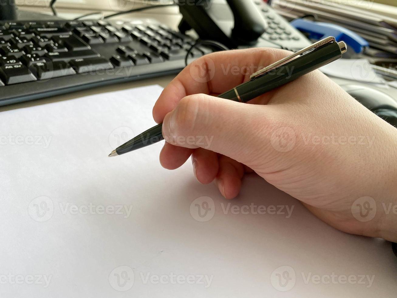 en mannens hand i en skjorta och med en kondition armband innehar en penna och skrivningar på de tabell på de kontor tabell med en dator med en tangentbord. företag arbete foto