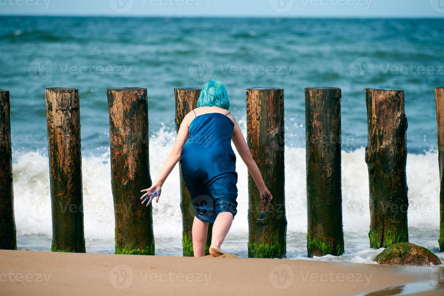 blåhårig kvinna performancekonstnär i blå klänning stående på stranden med pensel, bakifrån foto