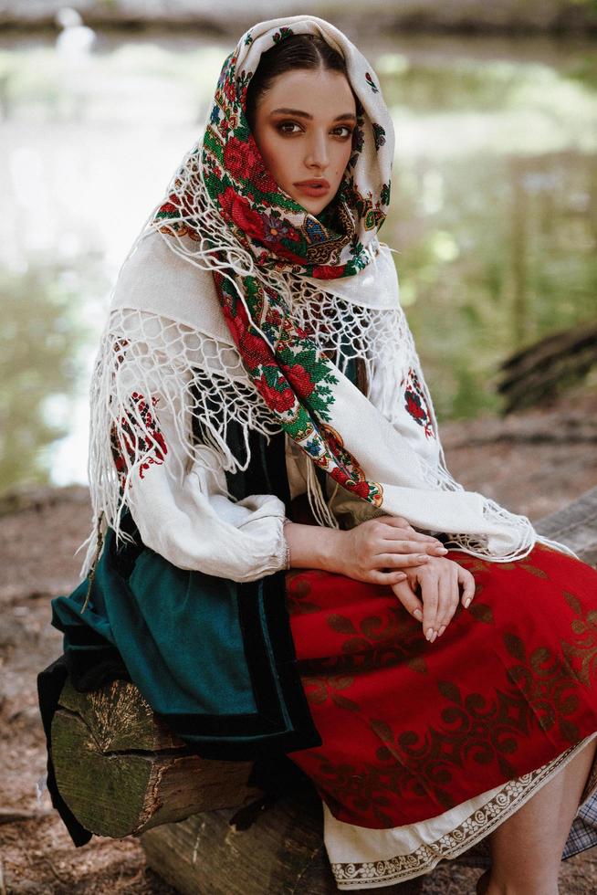 ung flicka i en traditionell broderad klänning som sitter på en bänk nära sjön foto