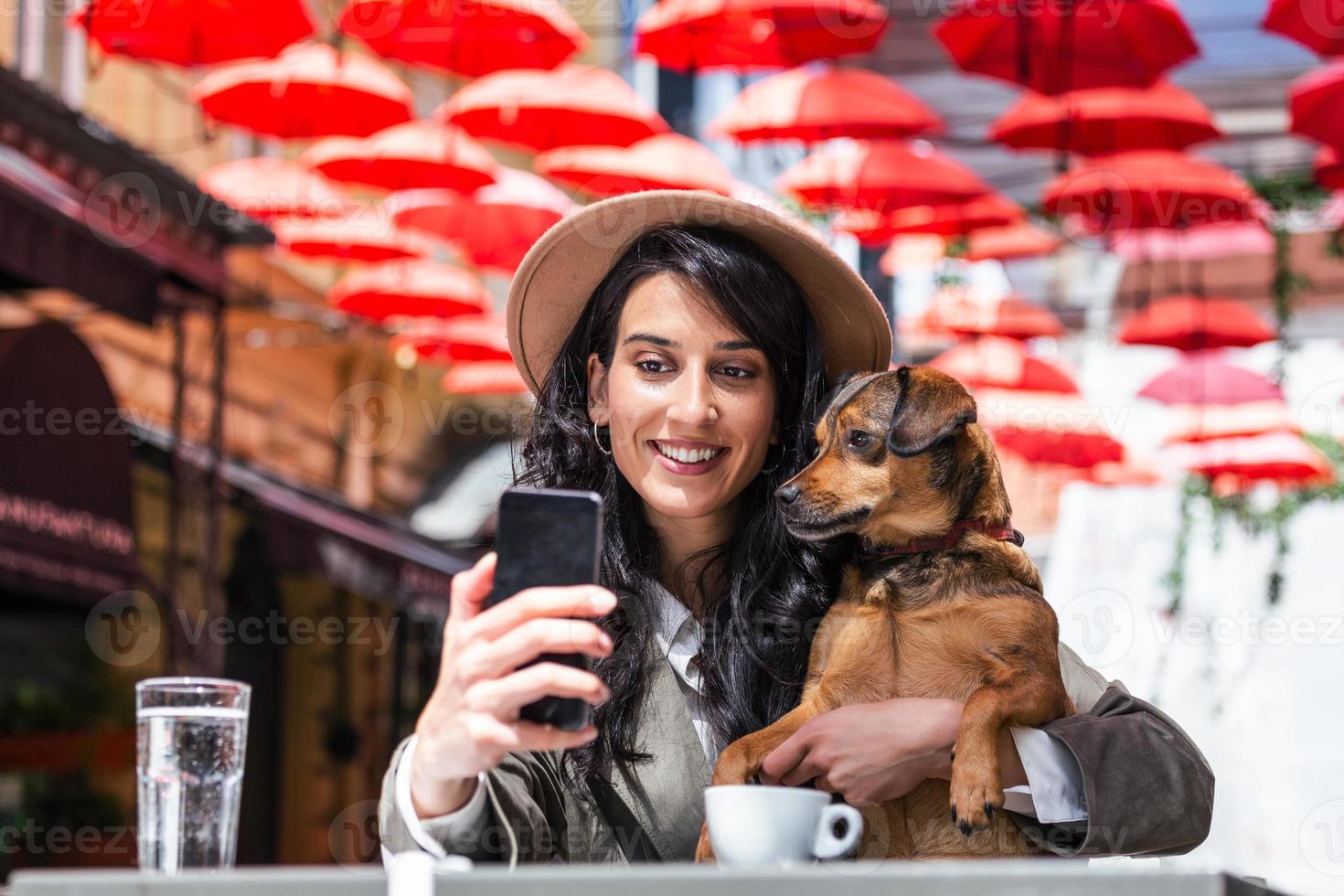 ung kvinna med henne hund i sällskapsdjur vänlig cafeteria. flicka dricka coffe på lokal- kaffe affär med henne sällskapsdjur hund tar en selfie med mobil telefon foto