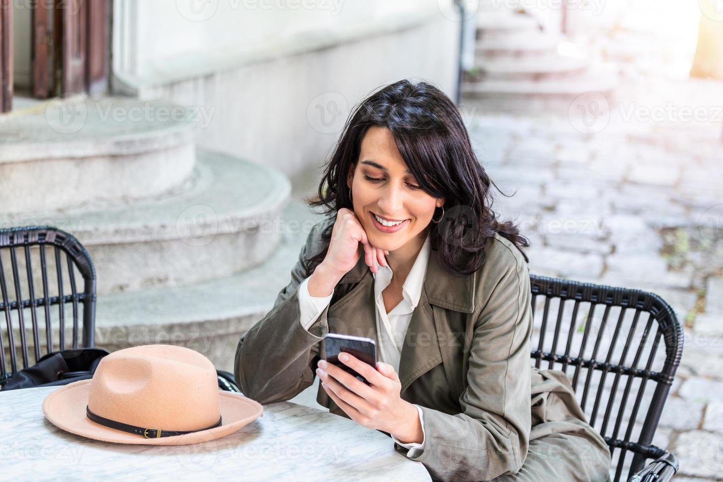 ung skön kvinna ser på smartphone medan Sammanträde på cafeteria. Lycklig universitet studerande använder sig av mobil telefon. affärskvinna dricka kaffe, leende och använder sig av smartphone inomhus. foto