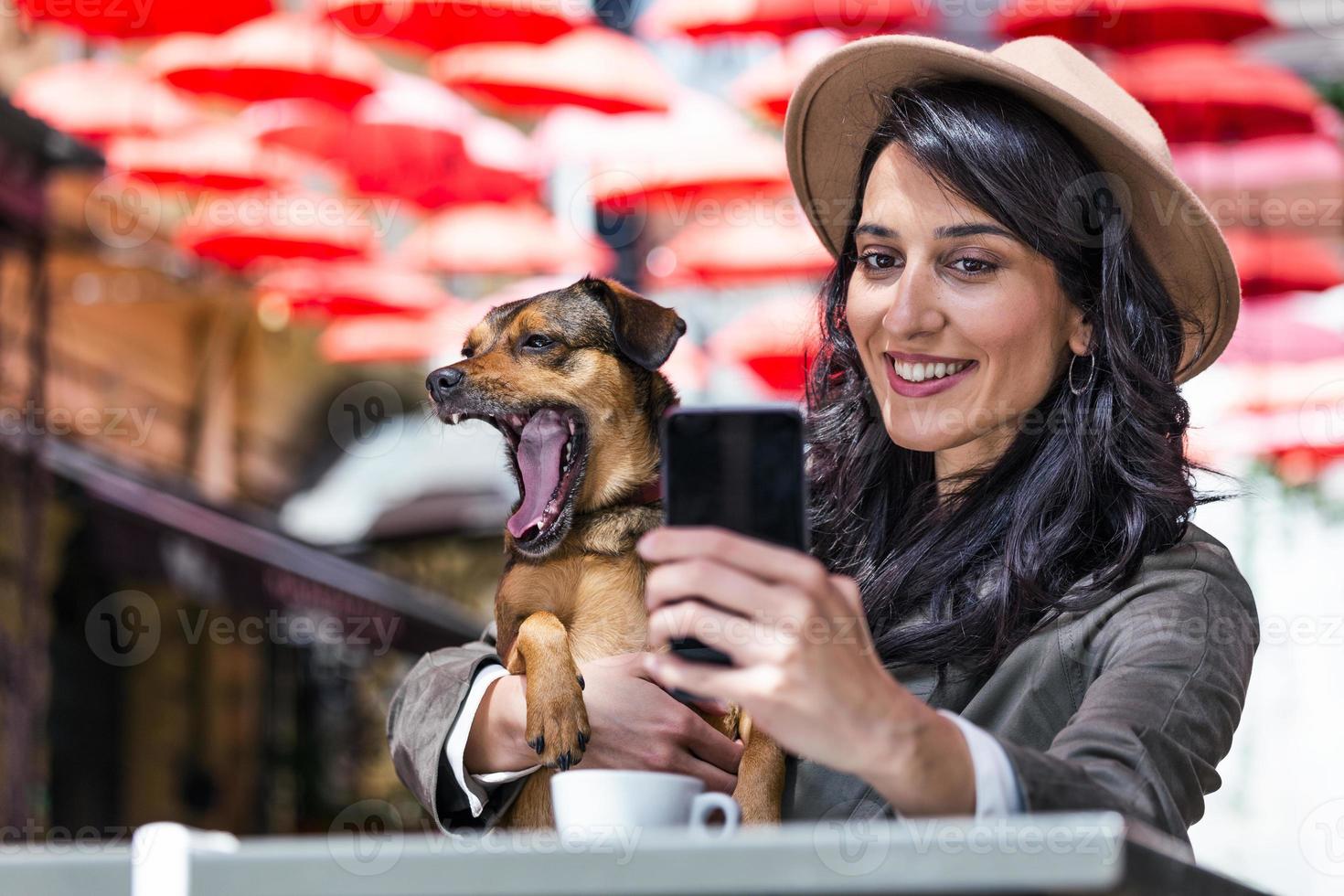 ung kvinna med henne hund i sällskapsdjur vänlig cafeteria. flicka dricka coffe på lokal- kaffe affär med henne sällskapsdjur hund tar en selfie med mobil telefon foto