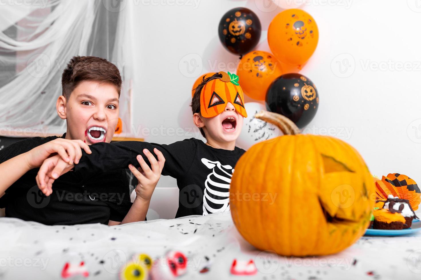 skrämmande pojke med vampyr huggtänder l låtsas till bita hans bröder ärm på de halloween fest. domkraft o' lykta halloween pumpa på de tabell foto