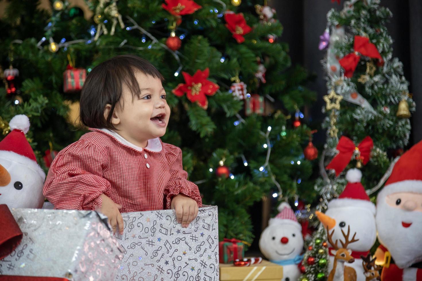 caucasian litet barn bebis är lyckligt leende medan har roligt spelar runt om gåva låda från föräldrar medan klä på sig i jul klänning med jul träd på de tillbaka för säsong firande begrepp foto