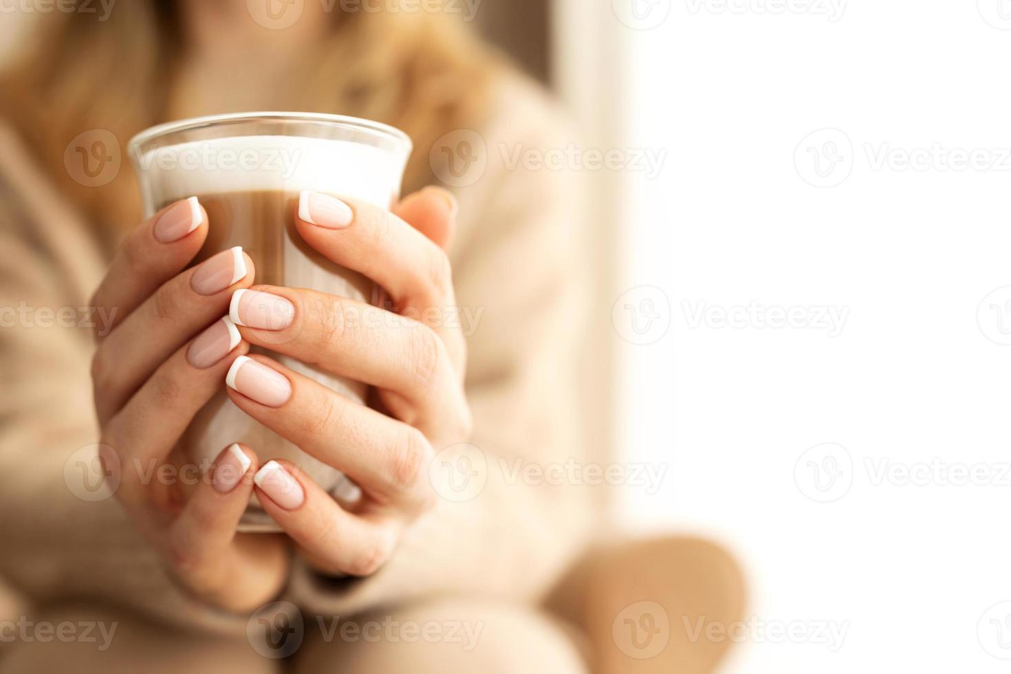 stänga upp kvinnors händer och fingrar med en skön franska manikyr innehav kopp transparent glas med kaffe med vit skum.kopia Plats för text, baner. beige stickat Tröja och bakgrund. foto