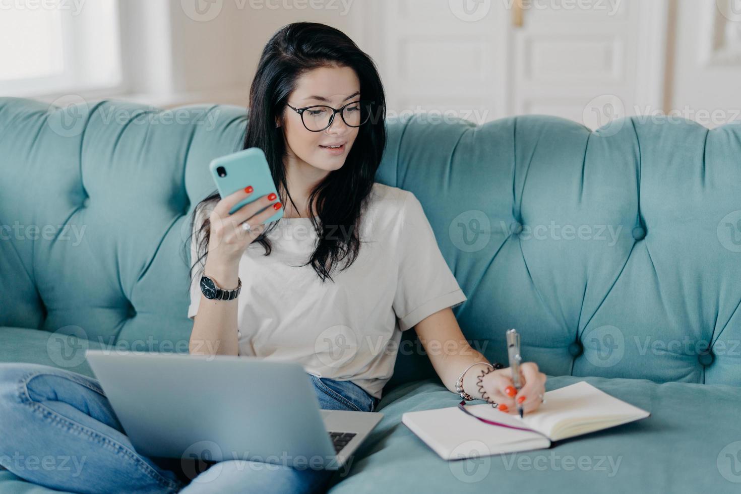 inomhus skott av söt brunett kvinnlig student tittar på webbseminarium och gör anteckningar i anteckningsblock, håller mobiltelefon, sitter i bekväm soffa, upptagen med distansjobb. människor, teknik, livsstilskoncept foto